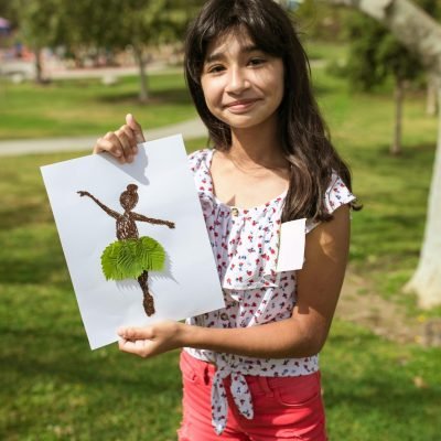 A young girl proudly shows her nature-themed artwork in a sunny park. Perfect for summer camp vibes.